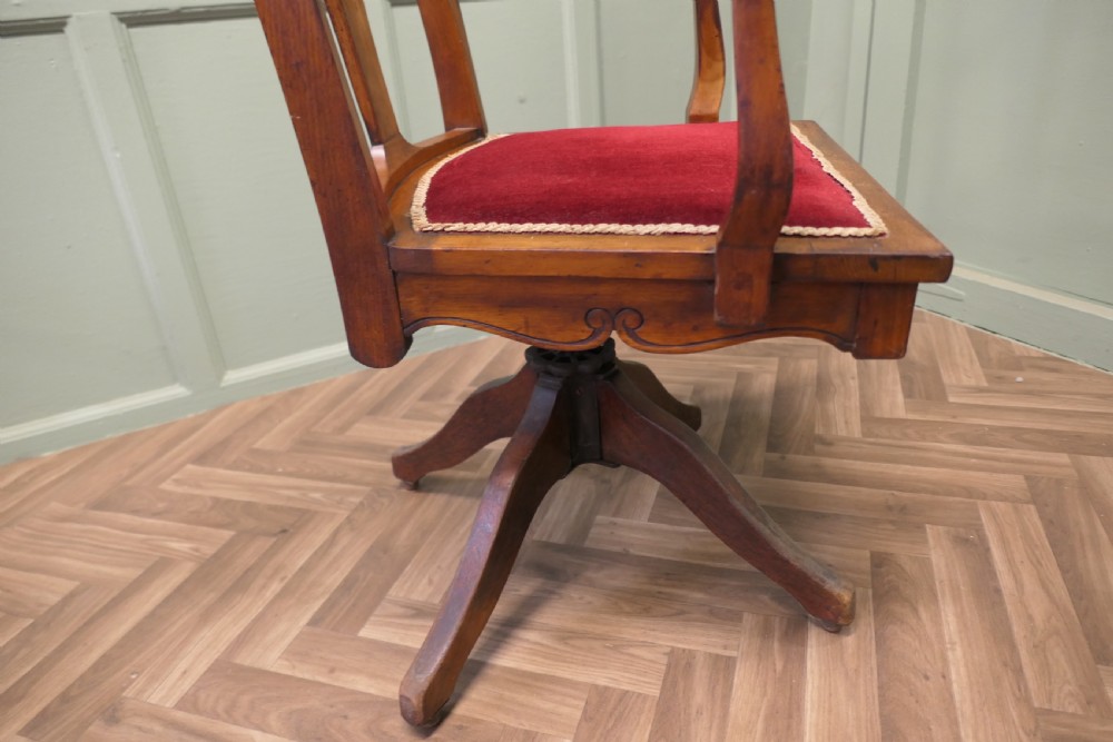 An Edwardian Arts and Crafts walnut revolving desk chair with a curved backrest, carved splat detail, shaped armrests, and a red velvet seat insert. View VI
