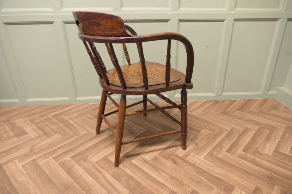 An antique oak smokers bow armchair featuring turned spindles, a curved top crest rail with a cut-out handle, and a perforated wooden seat panel. View V