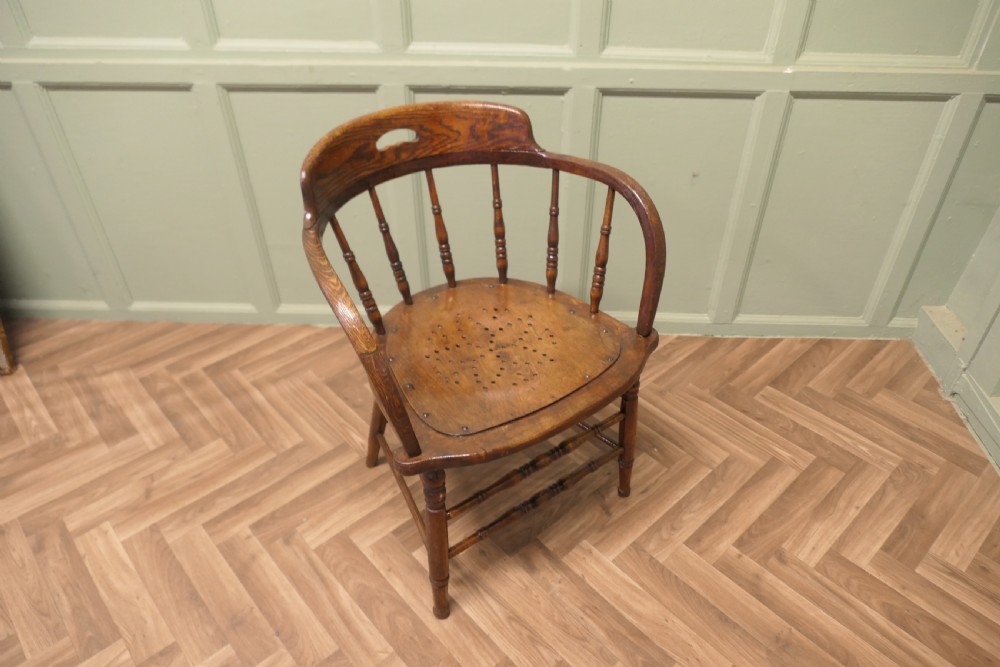 An antique oak smokers bow armchair featuring turned spindles, a curved top crest rail with a cut-out handle, and a perforated wooden seat panel. View VI
