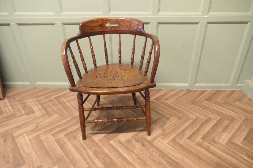 An antique oak smokers bow armchair featuring turned spindles, a curved top crest rail with a cut-out handle, and a perforated wooden seat panel. View VII