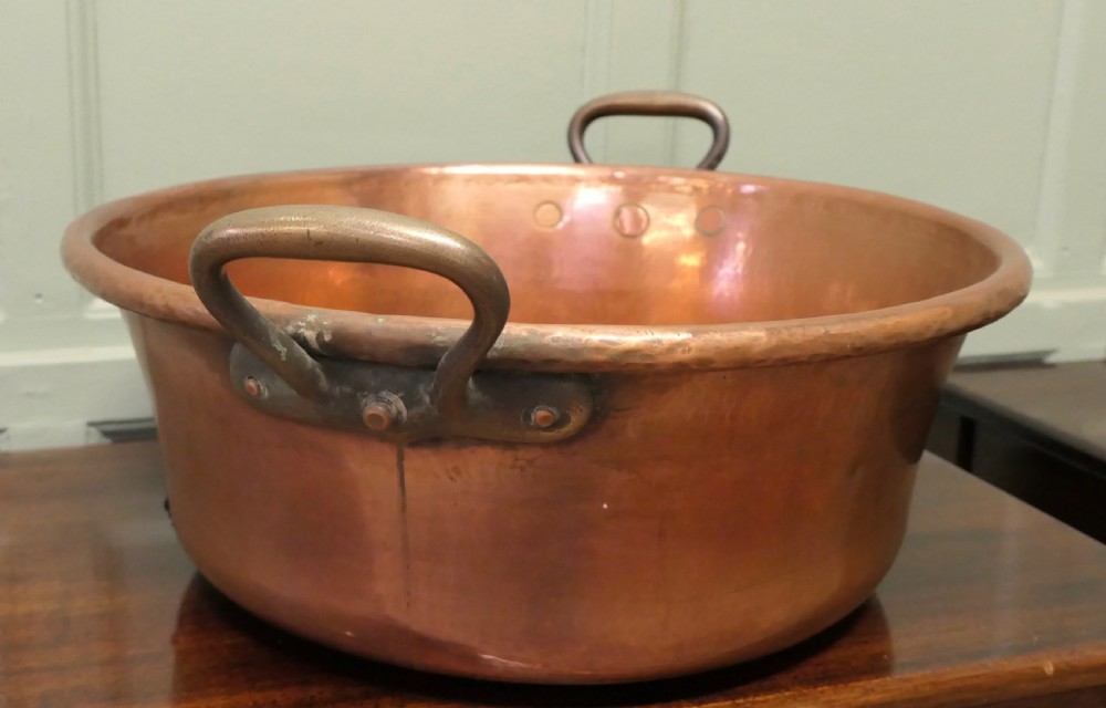 A large nineteenth-century hand-hammered copper jam pan with two cast side handles resting on a dark wood surface. View V