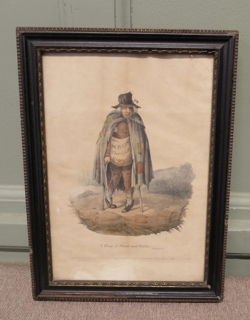 An early nineteenth-century framed hand-coloured print depicting a beggar figure with crutches and a patched cloak, titled A Thing of Shreds and Patches. View V