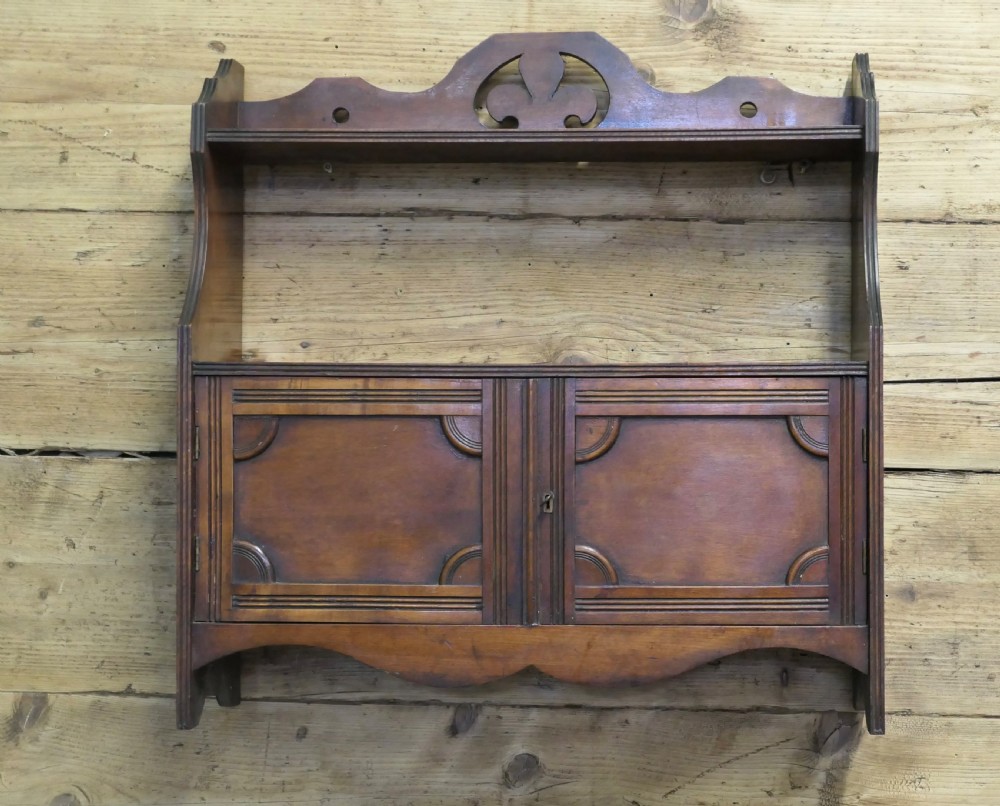 A warm brown wooden Art Deco wall cabinet featuring an upper open shelf with a pierced motif top rail above a two-door enclosed cupboard.