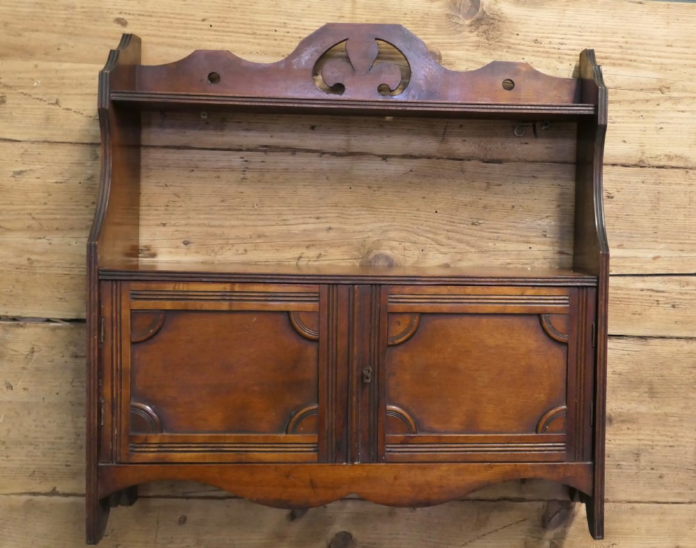 A warm brown wooden Art Deco wall cabinet featuring an upper open shelf with a pierced motif top rail above a two-door enclosed cupboard. View III