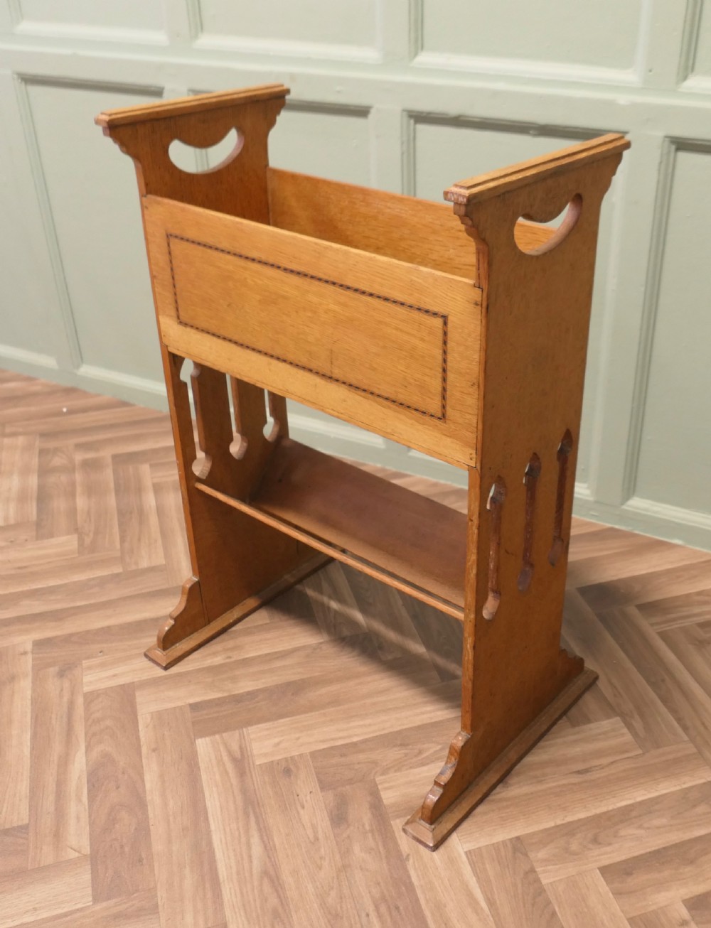 An Arts and Crafts golden oak two-tier bookcase featuring a upper trough with inlaid detail, side cut-out handles, and a lower shelf. View V