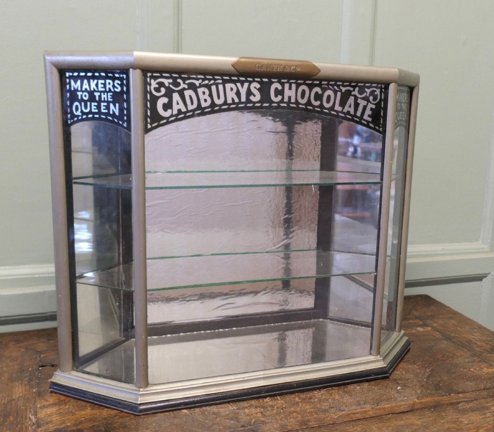 An Art Deco three-sided glass shop counter display cabinet with nickel-plated metal framing, glass shelves, mirrored back, and branded glass lettering reading Cadburys Chocolate Makers to the Queen.
