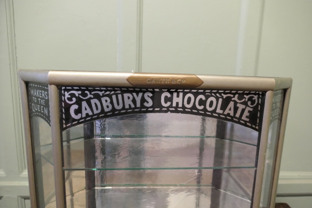 An Art Deco three-sided glass shop counter display cabinet with nickel-plated metal framing, glass shelves, mirrored back, and branded glass lettering reading Cadburys Chocolate Makers to the Queen. View II