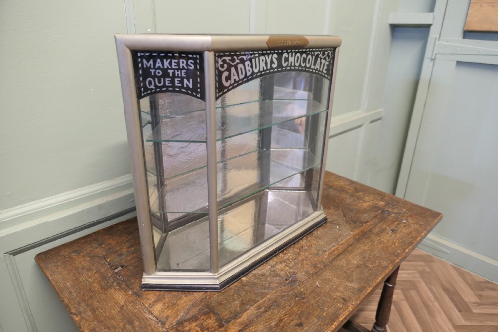 An Art Deco three-sided glass shop counter display cabinet with nickel-plated metal framing, glass shelves, mirrored back, and branded glass lettering reading Cadburys Chocolate Makers to the Queen. View IV