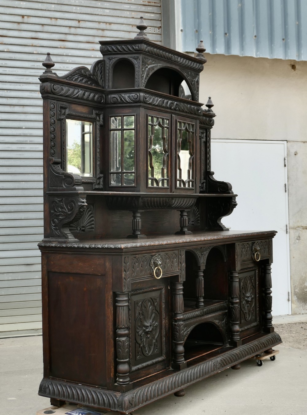 A large dark oak Victorian sideboard featuring deep Green Man carvings, a mirrored back superstructure, turned support columns, and lion head drawer handles.