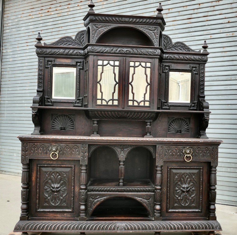 A large dark oak Victorian sideboard featuring deep Green Man carvings, a mirrored back superstructure, turned support columns, and lion head drawer handles. View II