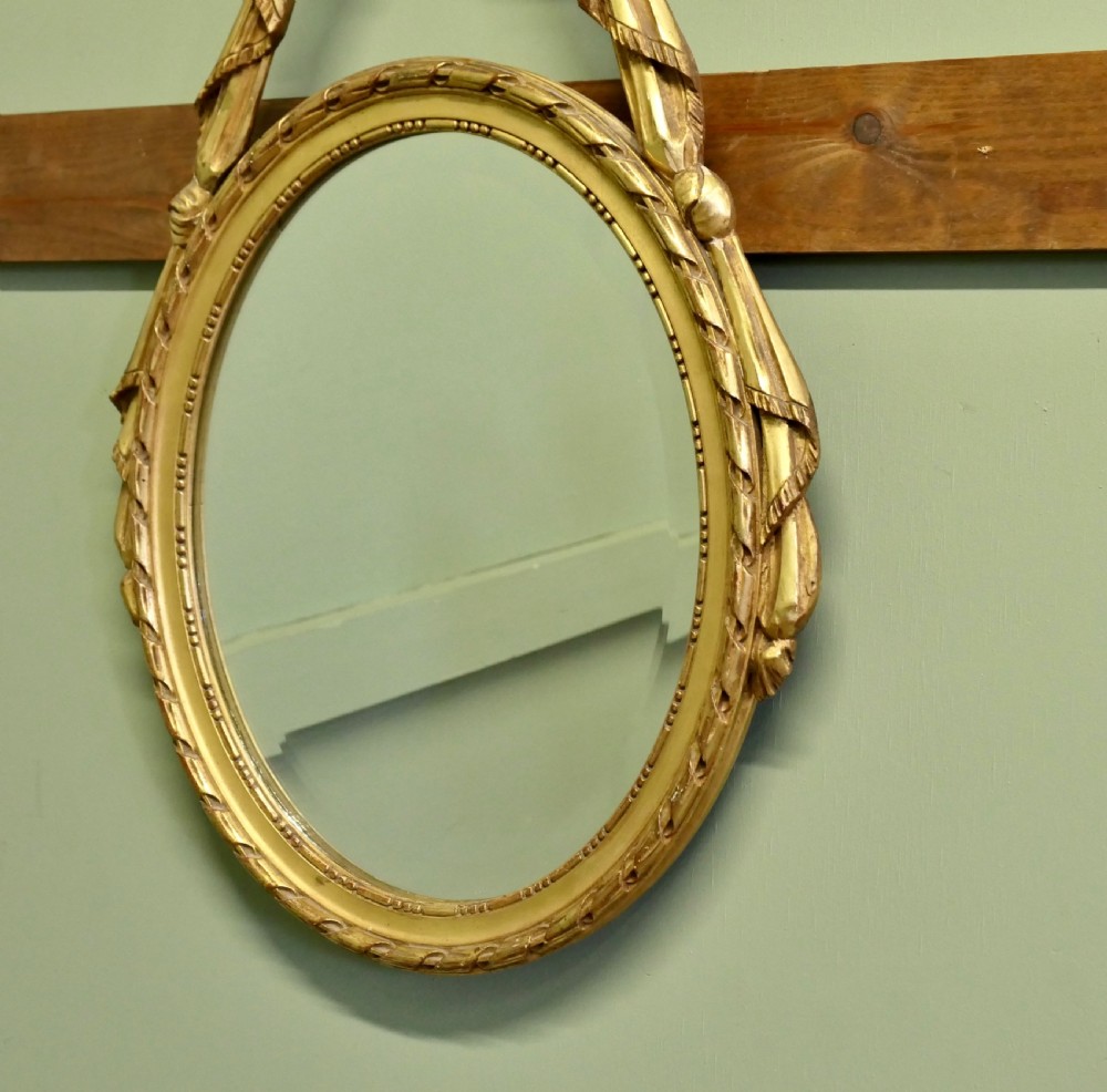 An oval wall mirror featuring a gold giltwood frame topped with a carved ribbon bow, torch, and quiver crest with trailing side ribbons. View II