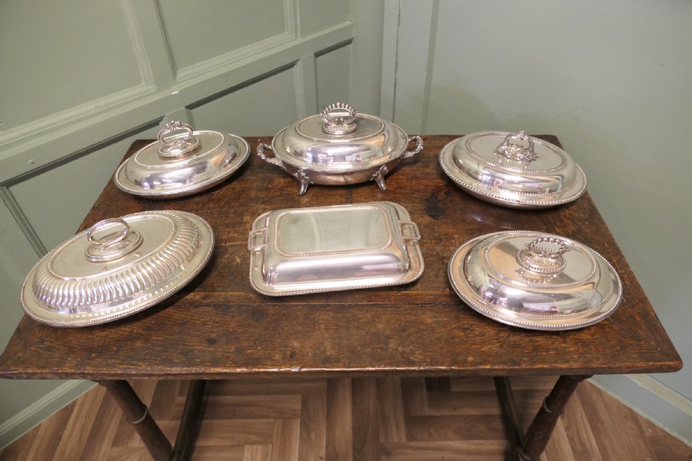 A six-piece collection of antique silver-plated serving tableware including oval entree dishes with covers, a footed tureen, and a rectangular tray arranged on a dark oak table.