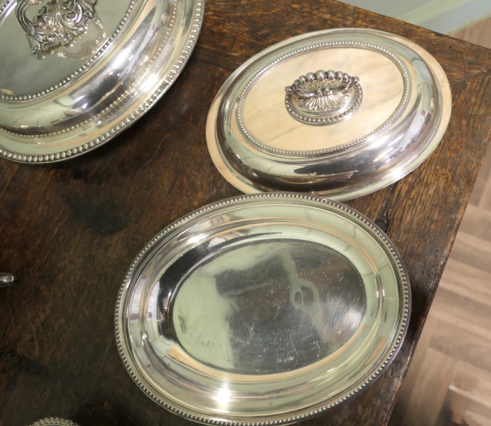 A six-piece collection of antique silver-plated serving tableware including oval entree dishes with covers, a footed tureen, and a rectangular tray arranged on a dark oak table. View II