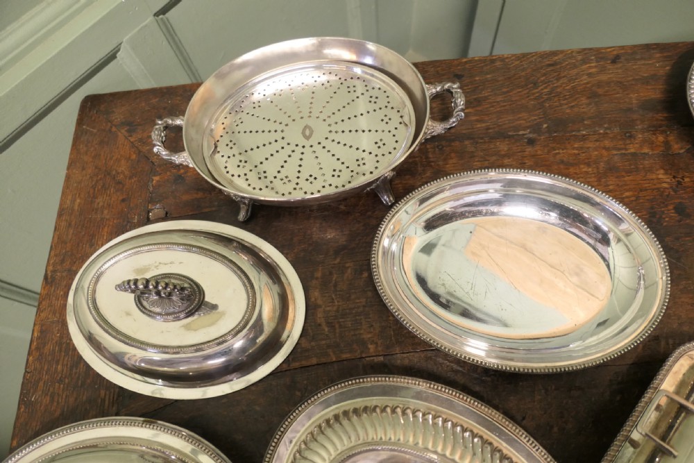 A six-piece collection of antique silver-plated serving tableware including oval entree dishes with covers, a footed tureen, and a rectangular tray arranged on a dark oak table. View III