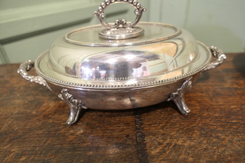 A six-piece collection of antique silver-plated serving tableware including oval entree dishes with covers, a footed tureen, and a rectangular tray arranged on a dark oak table. View IV