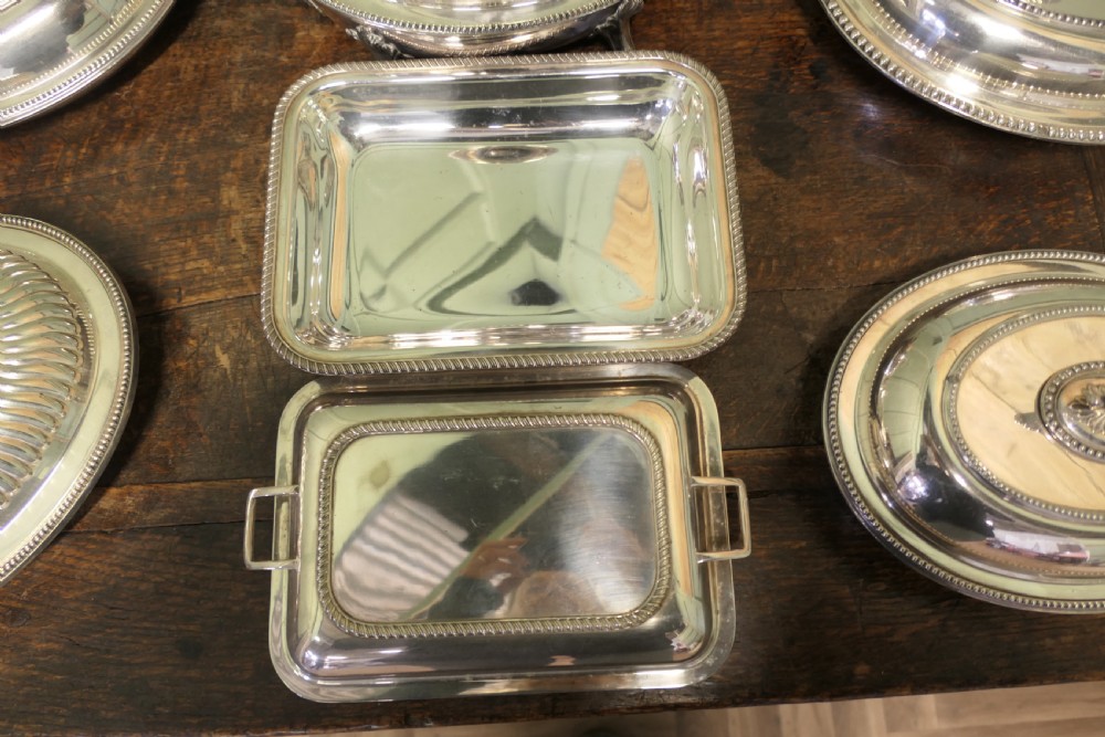 A six-piece collection of antique silver-plated serving tableware including oval entree dishes with covers, a footed tureen, and a rectangular tray arranged on a dark oak table. View V