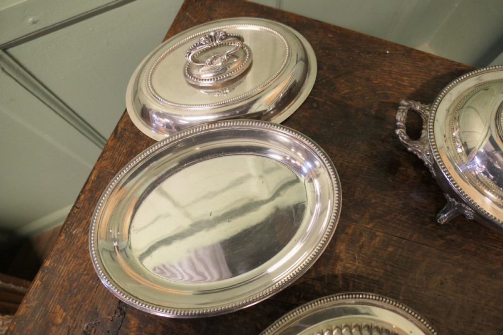 A six-piece collection of antique silver-plated serving tableware including oval entree dishes with covers, a footed tureen, and a rectangular tray arranged on a dark oak table. View VI