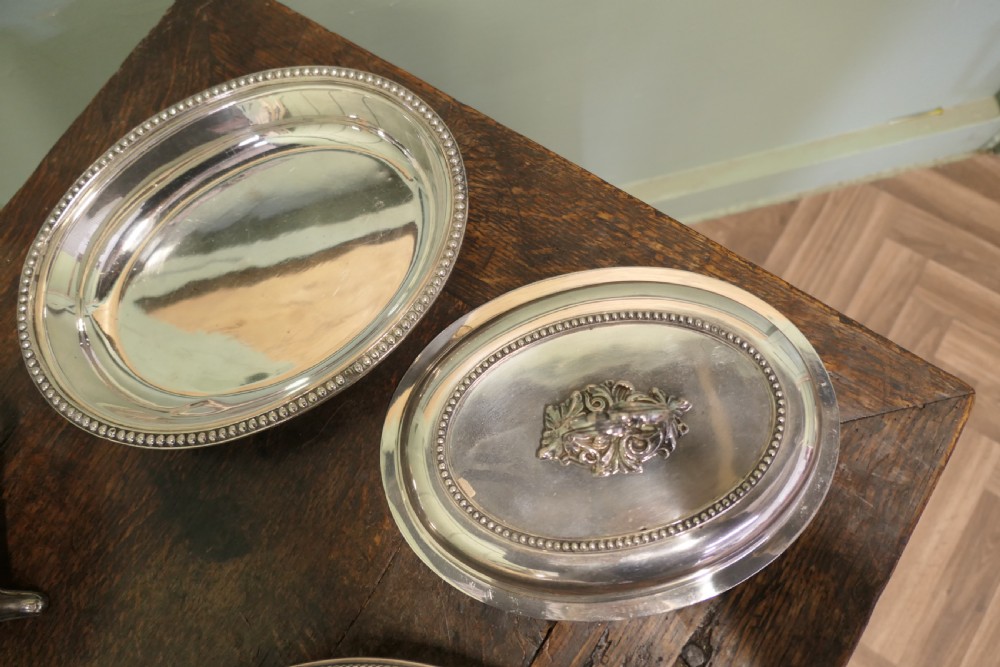 A six-piece collection of antique silver-plated serving tableware including oval entree dishes with covers, a footed tureen, and a rectangular tray arranged on a dark oak table. View VII