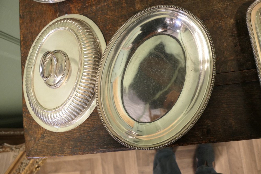 A six-piece collection of antique silver-plated serving tableware including oval entree dishes with covers, a footed tureen, and a rectangular tray arranged on a dark oak table. View VIII