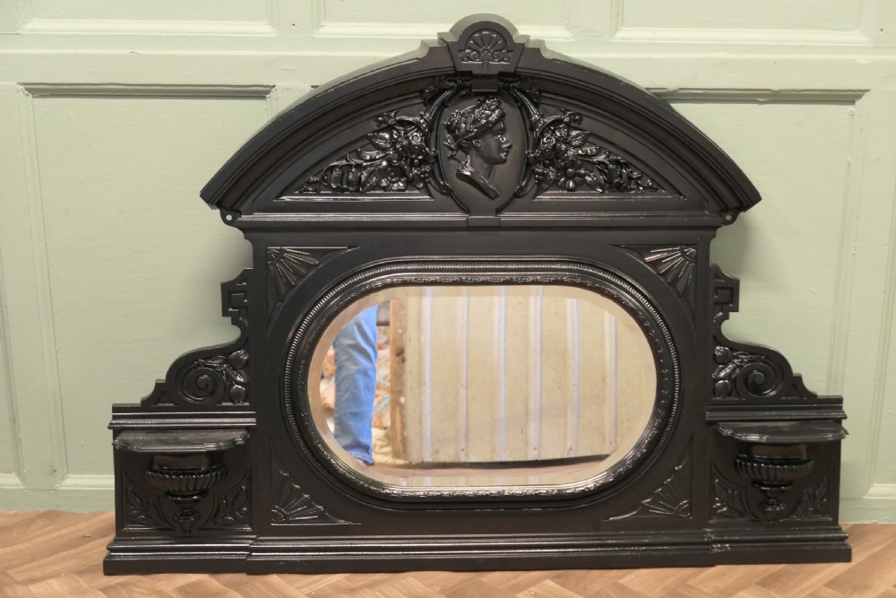 A black Victorian cast iron overmantel mirror featuring an arched pediment with a classical portrait medallion and an oval central mirror glass.