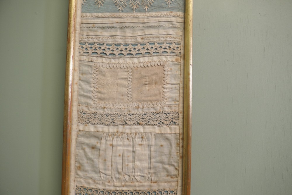 A tall, narrow gilt-framed vertical sampler displaying multiple horizontal bands of intricate white lace, pleating, and embroidered text over a pale blue background. View II