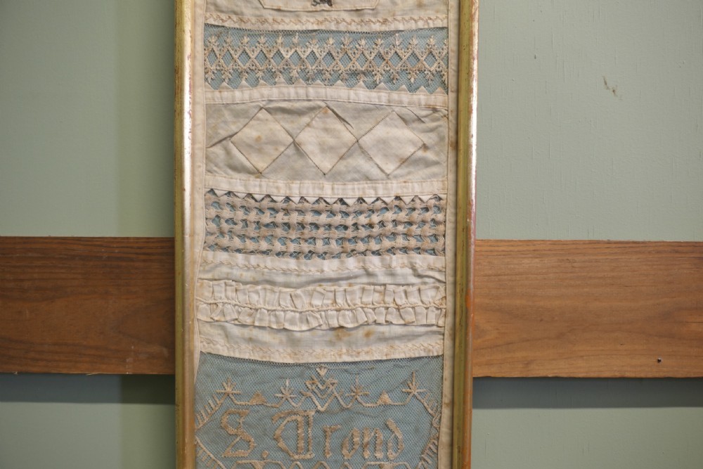 A tall, narrow gilt-framed vertical sampler displaying multiple horizontal bands of intricate white lace, pleating, and embroidered text over a pale blue background. View III
