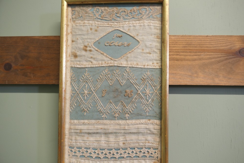 A tall, narrow gilt-framed vertical sampler displaying multiple horizontal bands of intricate white lace, pleating, and embroidered text over a pale blue background. View IV