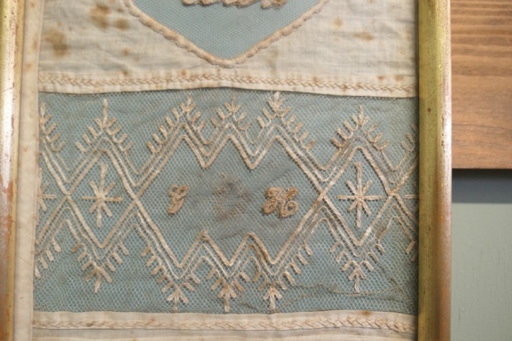 A tall, narrow gilt-framed vertical sampler displaying multiple horizontal bands of intricate white lace, pleating, and embroidered text over a pale blue background. View V