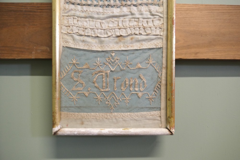 A tall, narrow gilt-framed vertical sampler displaying multiple horizontal bands of intricate white lace, pleating, and embroidered text over a pale blue background. View VI