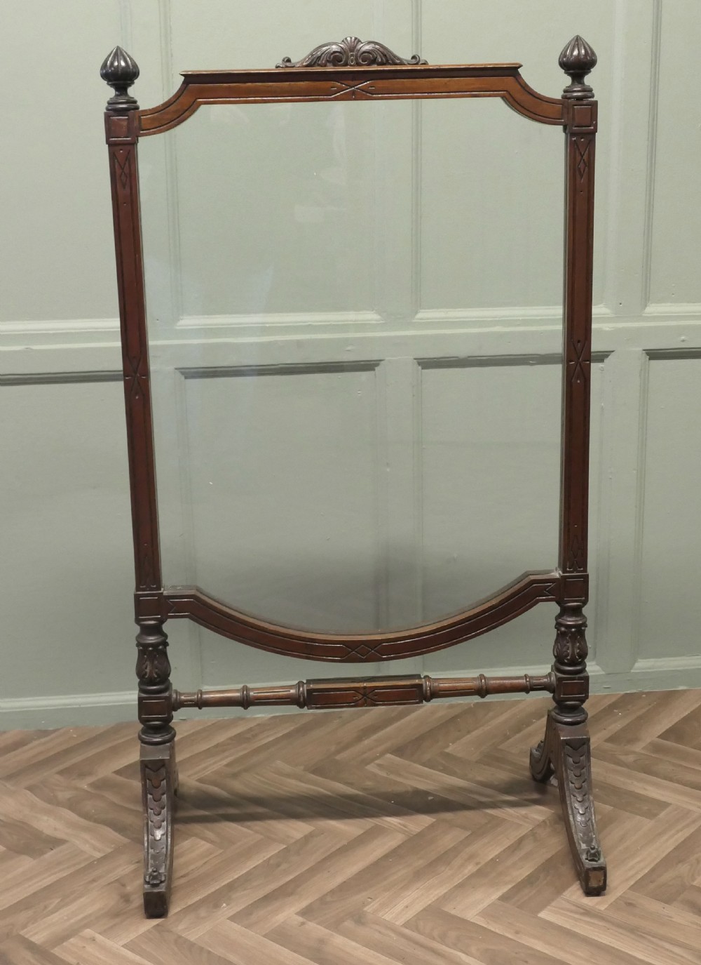 A freestanding Victorian dark mahogany fire screen featuring a clear glass panel, carved finials, turned side columns, and splayed trestle feet. View II