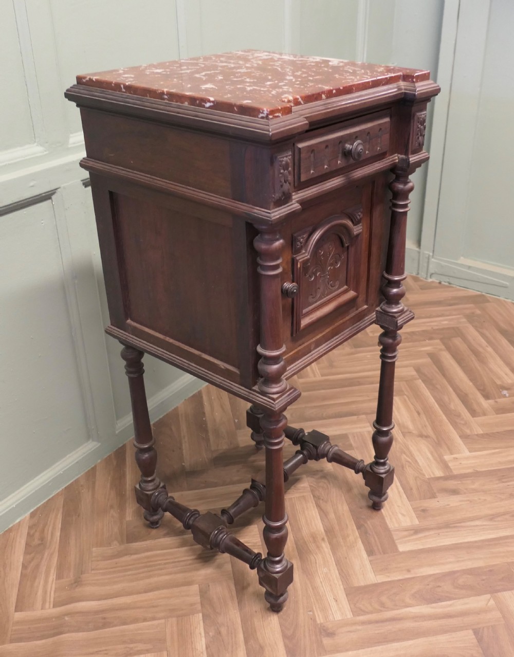 A dark carved wood French bedside table featuring a red marble top, single drawer, cupboard door with fleur-de-lis motif, and turned legs. View III