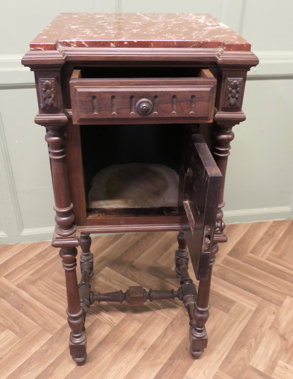 A dark carved wood French bedside table featuring a red marble top, single drawer, cupboard door with fleur-de-lis motif, and turned legs. View V