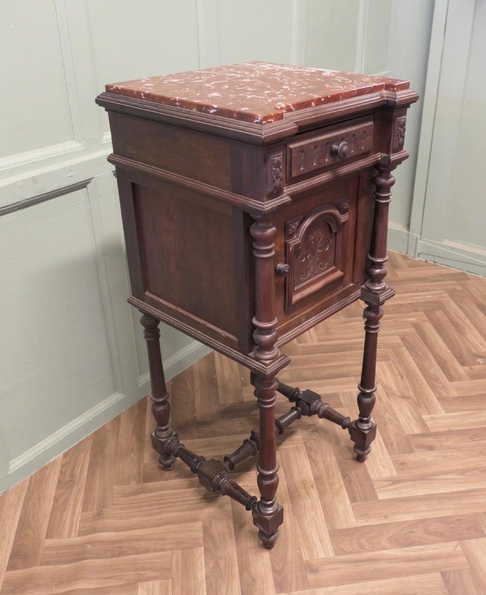 A dark carved wood French bedside table featuring a red marble top, single drawer, cupboard door with fleur-de-lis motif, and turned legs. View VI