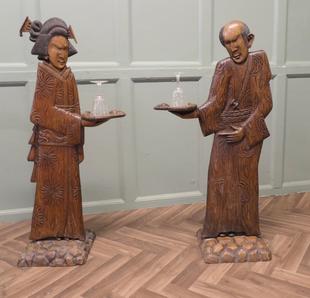 A pair of tall carved wood Japanese figures standing on stone-textured bases, each wearing detailed patterned kimonos and holding a raised tray.