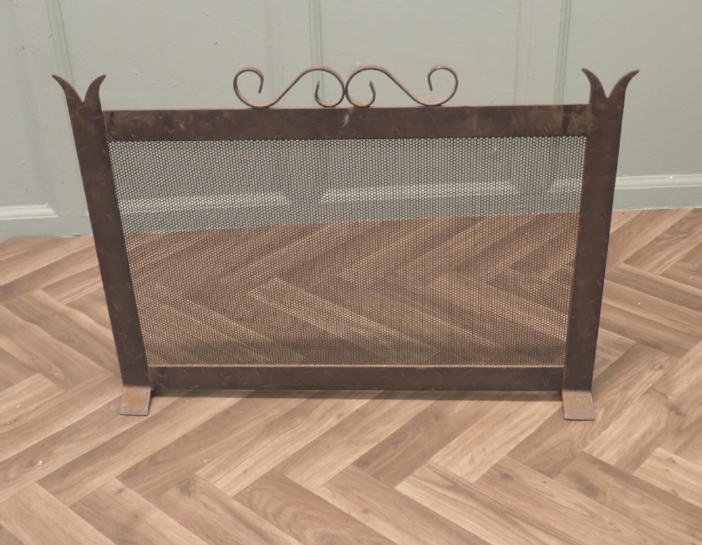 A dark wrought iron fire screen with a perforated mesh central panel, scrollwork top rail, and pointed upright posts resting on flat feet.