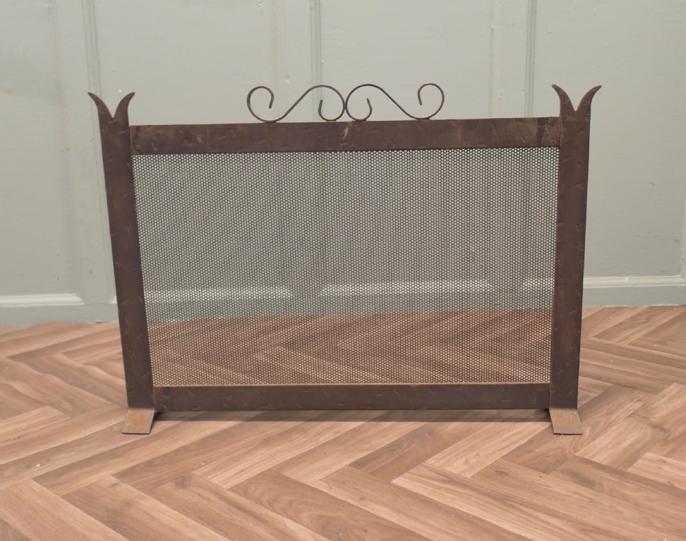 A dark wrought iron fire screen with a perforated mesh central panel, scrollwork top rail, and pointed upright posts resting on flat feet. View II