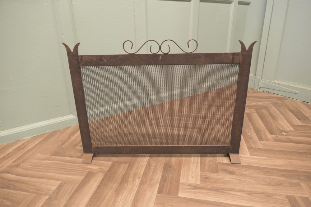 A dark wrought iron fire screen with a perforated mesh central panel, scrollwork top rail, and pointed upright posts resting on flat feet. View VI