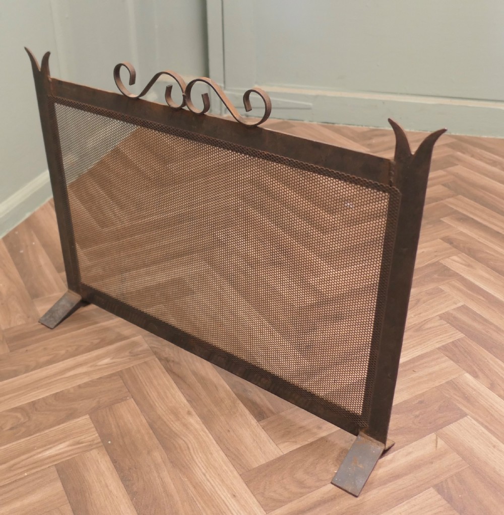 A dark wrought iron fire screen with a perforated mesh central panel, scrollwork top rail, and pointed upright posts resting on flat feet. View VII