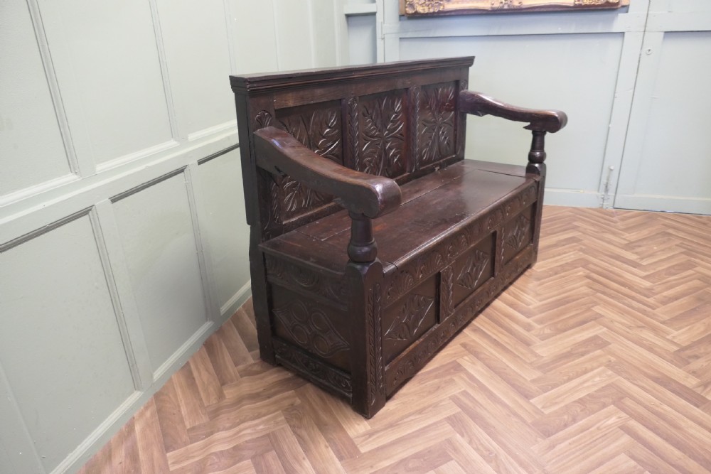 A dark carved oak high back box settle featuring three carved back panels, a hinged seat, and carved front base panels. View III