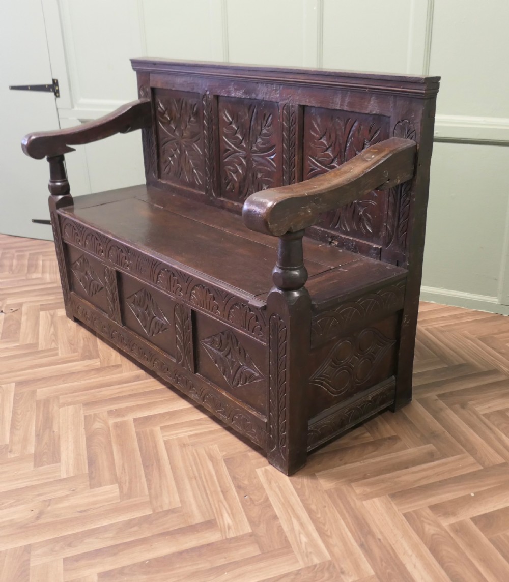 A dark carved oak high back box settle featuring three carved back panels, a hinged seat, and carved front base panels. View VI