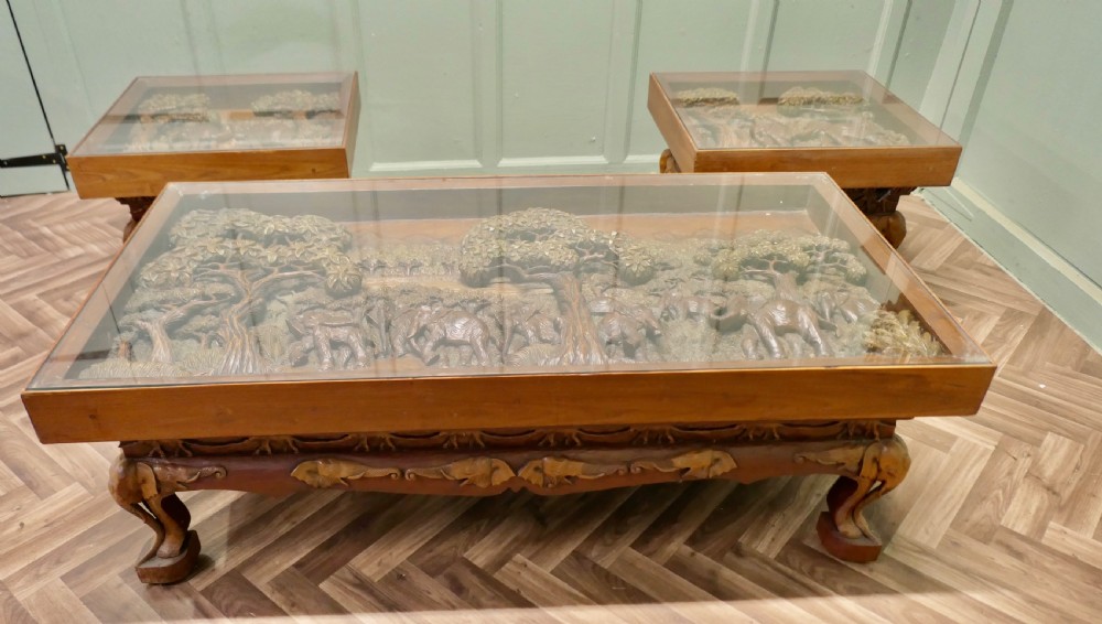A three-piece teak table set featuring elaborate relief carvings of elephants amidst trees under glass tops.