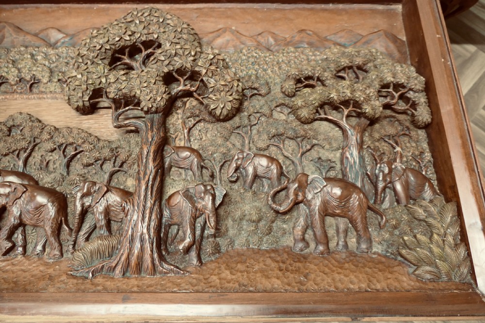 A three-piece teak table set featuring elaborate relief carvings of elephants amidst trees under glass tops. View III