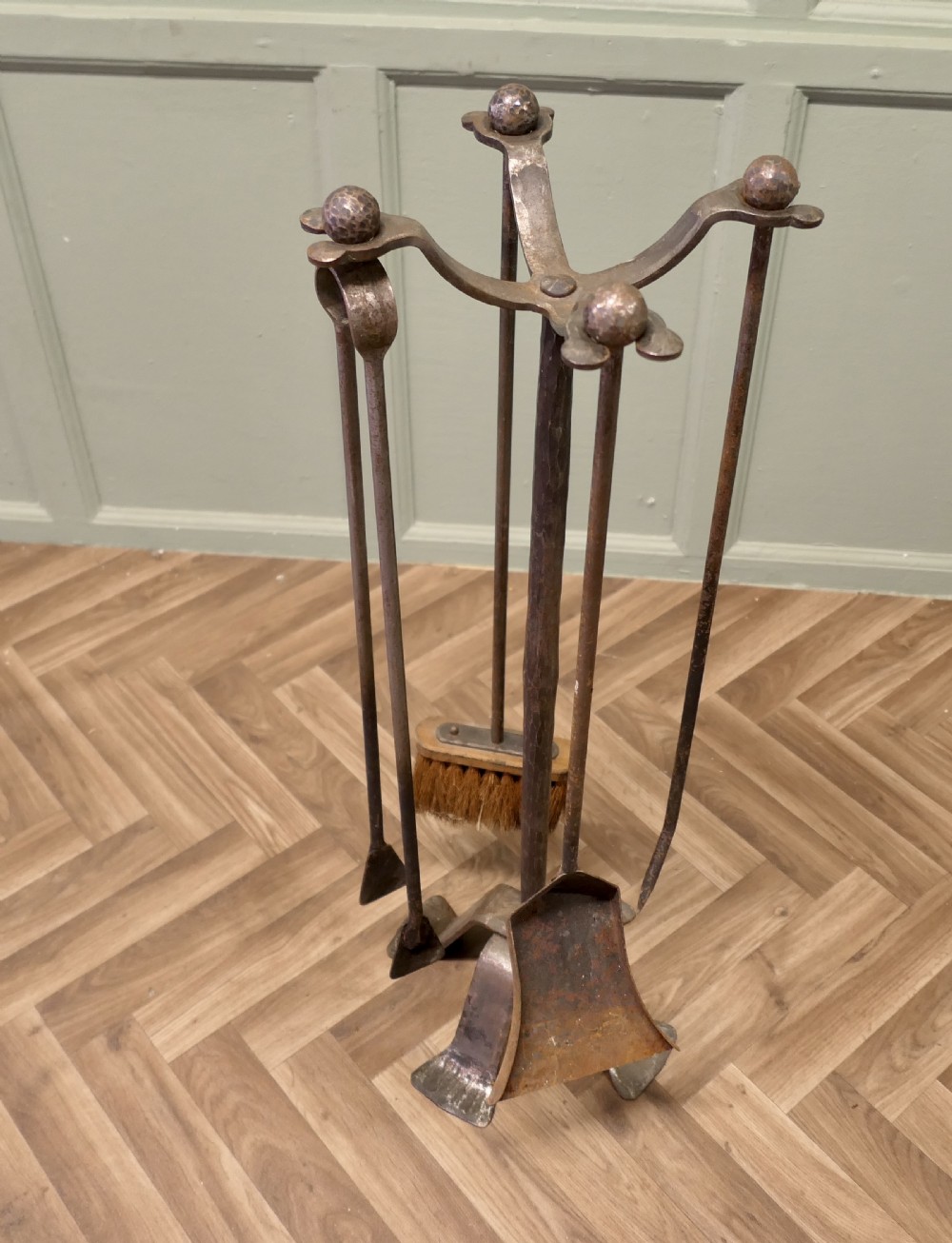 A dark hand-forged wrought iron fireplace companion set featuring a central stand with curved hooks holding a poker, shovel, tongs, and wooden brush. View VI