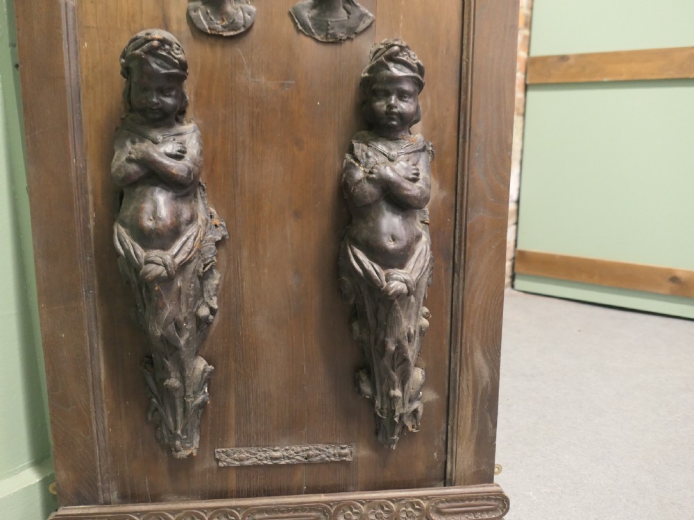 A pair of moulded dark brown leathermache putti term figures with arms crossed, mounted vertically on a timber panel.