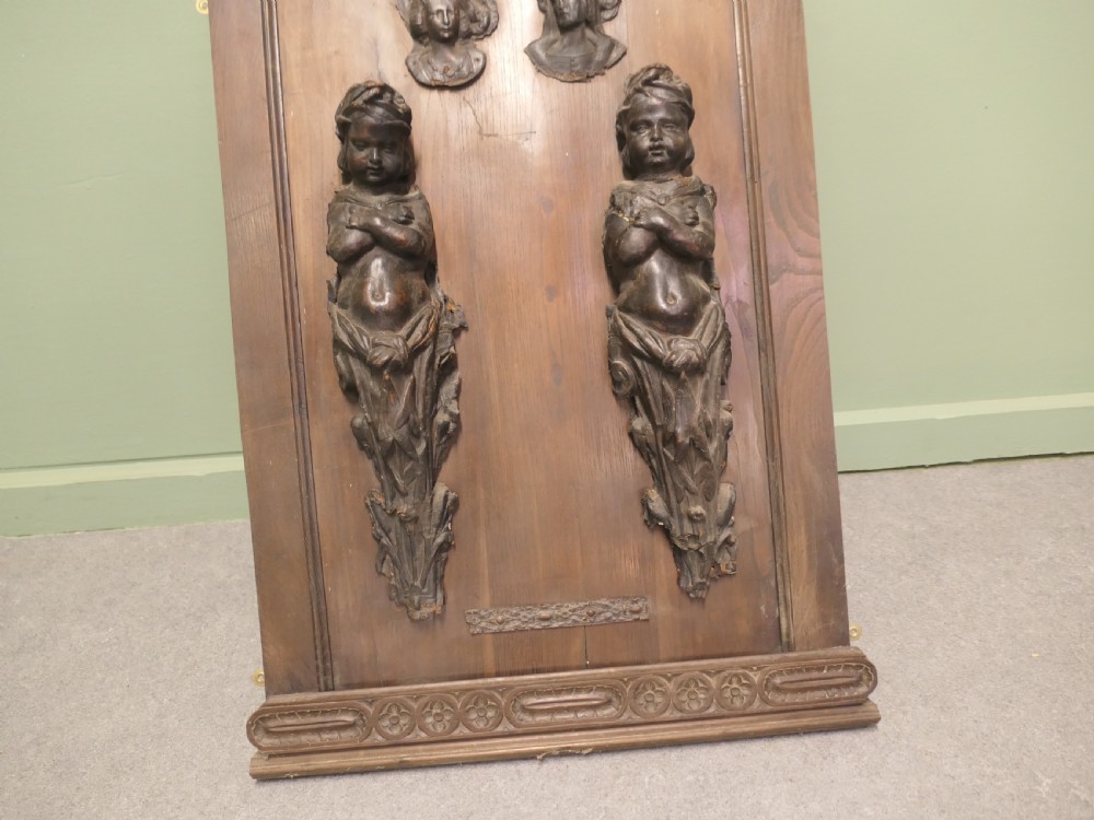 A pair of moulded dark brown leathermache putti term figures with arms crossed, mounted vertically on a timber panel. View II