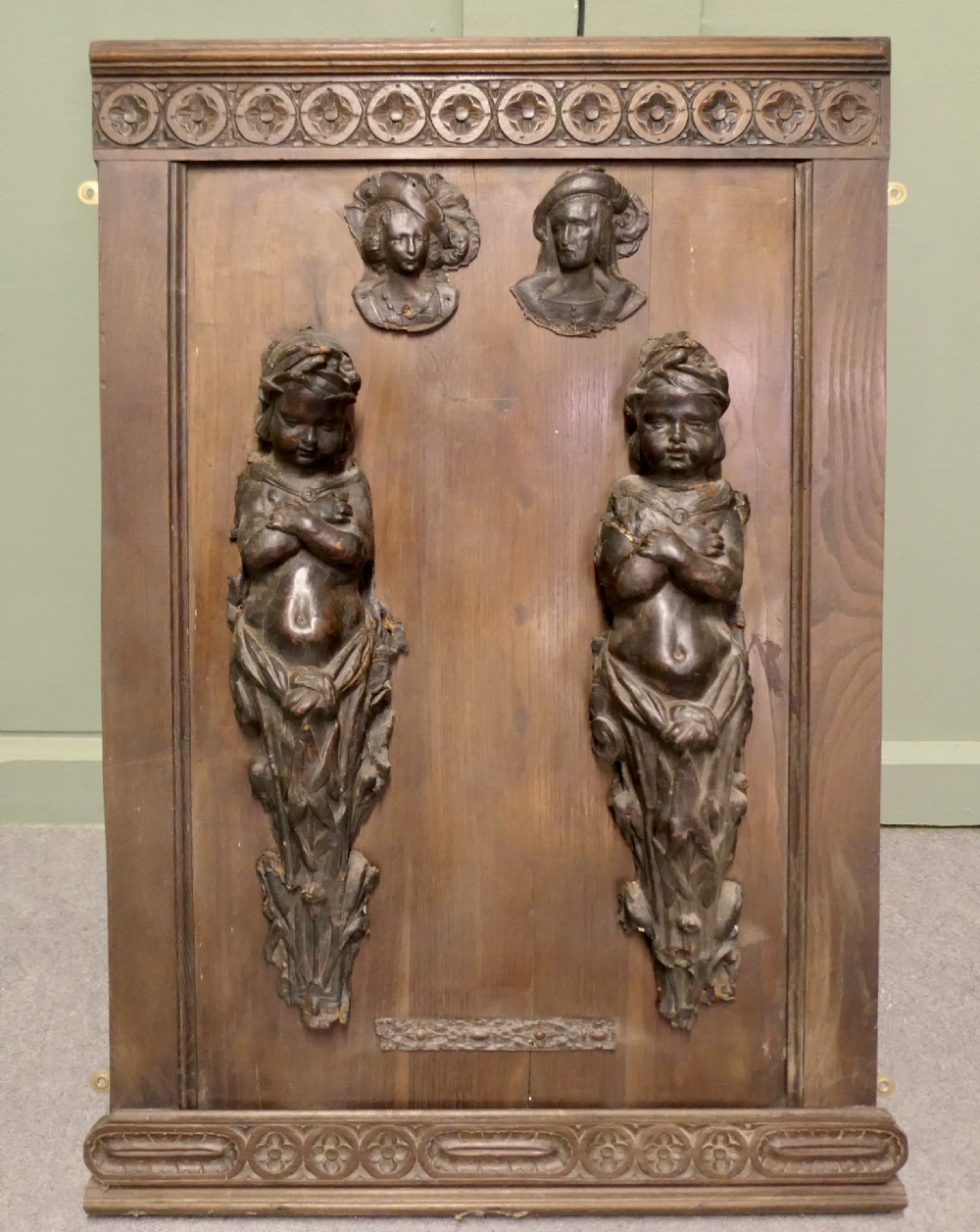 A pair of moulded dark brown leathermache putti term figures with arms crossed, mounted vertically on a timber panel. View III