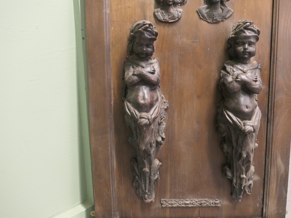 A pair of moulded dark brown leathermache putti term figures with arms crossed, mounted vertically on a timber panel. View V