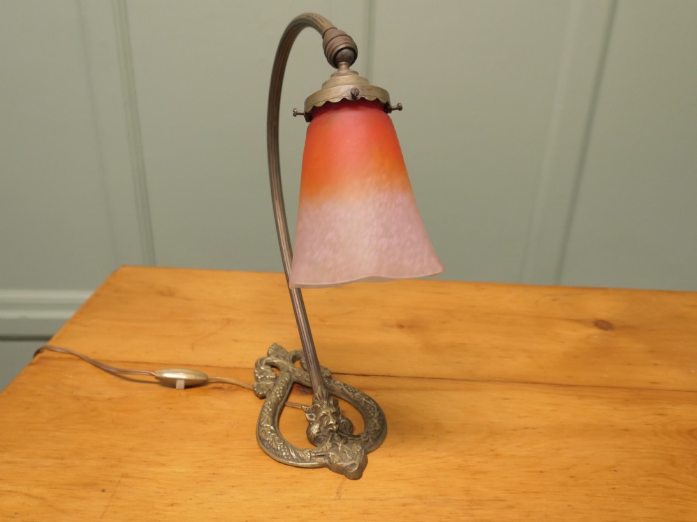 A French cast brass desk lamp featuring a curved gooseneck arm, an openwork base with a Bacchus face motif, and a flared frosted glass shade with an orange to pale pink gradient.