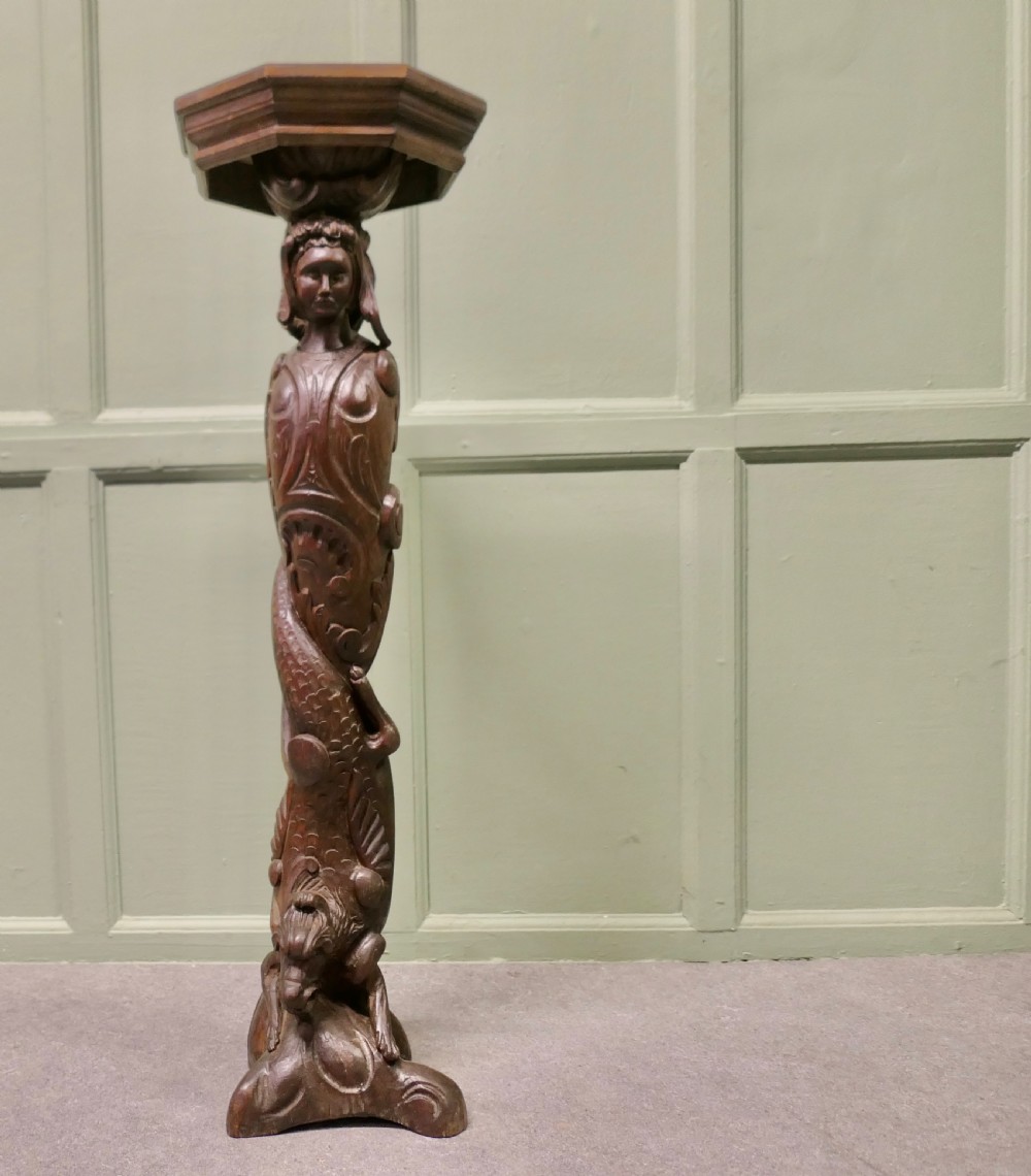 A dark carved oak pedestal featuring a caryatid figure and relief serpent design supporting an octagonal top platform.