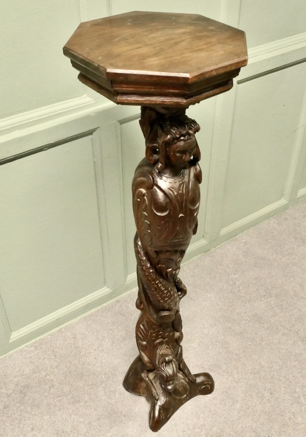A dark carved oak pedestal featuring a caryatid figure and relief serpent design supporting an octagonal top platform. View V